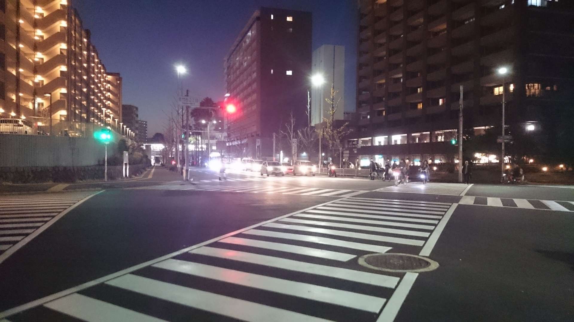 Kuvassa on yöaikaan valaistu kaupunkiristeys, jossa on selkeästi maalatut suojatiet. Taustalla näkyy korkeita kerrostaloja, joiden ikkunoista loistaa valoa, ja katuvalot valaisevat ympäristöä. Oikealla puolella on useita ihmisiä, jotka ylittävät tietä tai odottavat vuoroaan. Kuvassa näkyy myös liikennevaloja, joista osa näyttää punaista ja osa vihreää, mikä hallitsee risteyksen liikennettä. Ympäristö on tyypillinen moderni kaupunki-ilmapiiri.