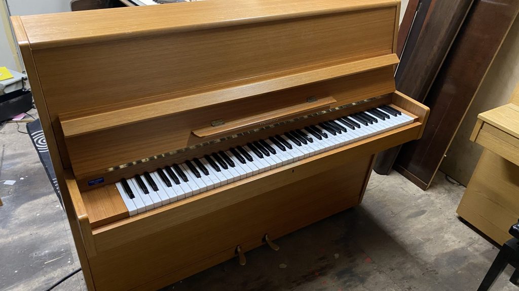 Vaaleasta puusta valmistettu pystypiano, jossa on minimalistinen ja käytännöllinen muotoilu. Koskettimisto on näkyvillä, ja pianon alareunassa on kaksi poljinta. Piano on sijoitettu sisätilaan, jossa näkyy myös muita huonekaluja ja soittimia.